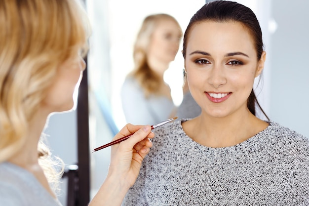 Professionelle Maskenbildnerin, die mit einer schönen jungen Frau arbeitet. Braut-, Mode- oder Nude-Stil. Beauty-Konzept.