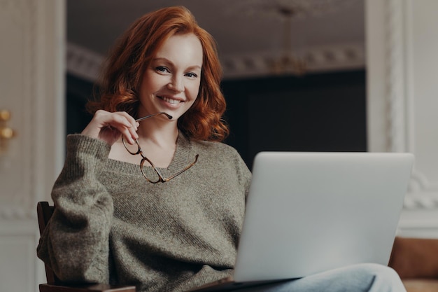 Professionelle positive rothaarige junge Europäerin arbeitet freiberuflich, konzentriert sich auf Remote-Job, bereitet Veröffentlichung für Webseite vor, sitzt vor geöffnetem Laptop-Computer und lächelt sanft