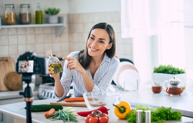 Professionelle schöne glückliche junge Frau bloggt für ihren Küchenkanal über gesundes Leben in der Küche ihres Hauses und schaut vor der Kamera auf ein Stativ