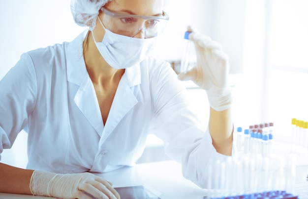 Professionelle Wissenschaftlerin in Schutzbrillen, die Röhren mit Reagenzien im sonnigen blau getönten Labor erforscht. Medizin und Wissenschaft forschen.