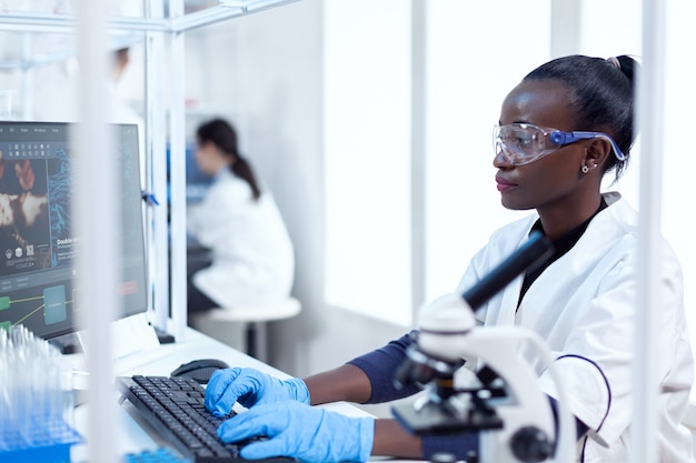 Professioneller afrikanischer Mediziner, der Informationen zur Virusstudie eingibt Schwarzer Gesundheitswissenschaftler im biochemischen Labor mit steriler Ausrüstung.