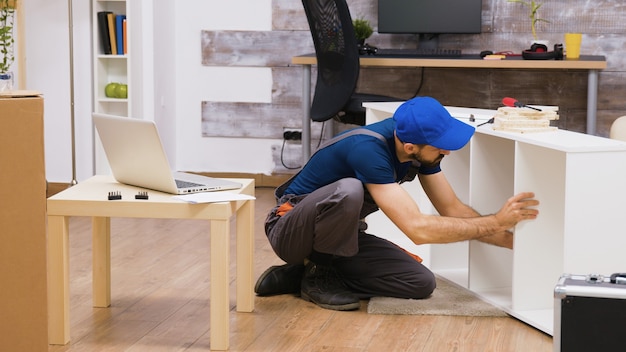 Professioneller Arbeiter in Overalls konsultiert Möbelmontageanleitungen vom Laptop. Arbeiter mit Mütze.