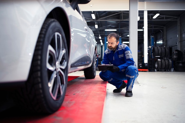 Professioneller bärtiger kaukasischer Automechaniker mittleren Alters, der eine Sichtprüfung des Fahrzeugs in der Werkstatt durchführt und eine Checkliste hält.