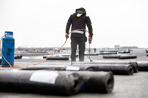 professioneller Dachträger, der auf einer modernen Baustelle mit einem Gasbrenner Bitumenrollen auf ein Flachdach aufträgt