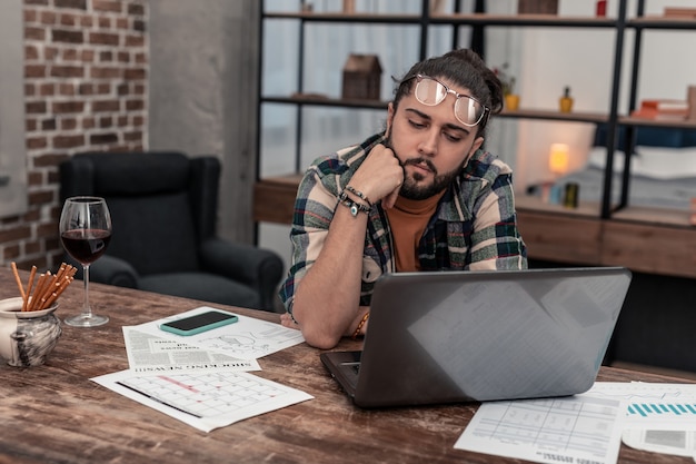 Professioneller Freiberufler. Angenehmer nachdenklicher Mann, der auf den Laptop-Bildschirm schaut, während er sich auf seine Arbeit konzentriert