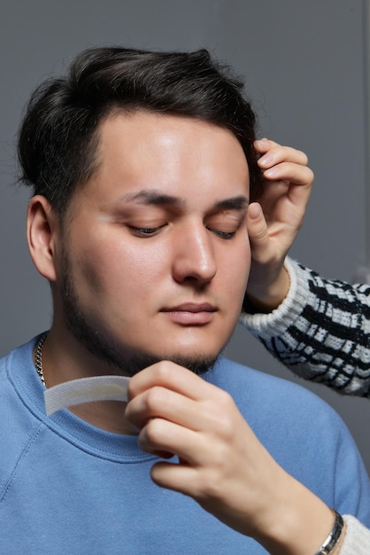 Professioneller Friseur, der das Einfädeln durchführt und die Form der Augenbrauen für junge männliche Kunden mit einer Pinzette im Friseursalon korrigiert Friseur bei der Arbeit Brauenpflegekonzept Brauen zupfen