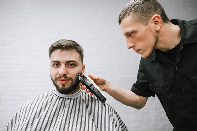 Professioneller Friseur schneidet einen positiven bärtigen Kunden mit einem Clipper gegen eine weiße Wand