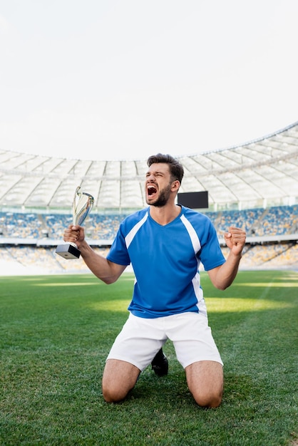 Professioneller Fußballspieler in blau-weißer Uniform mit Sportpokal, der auf Knien auf Fußball steht