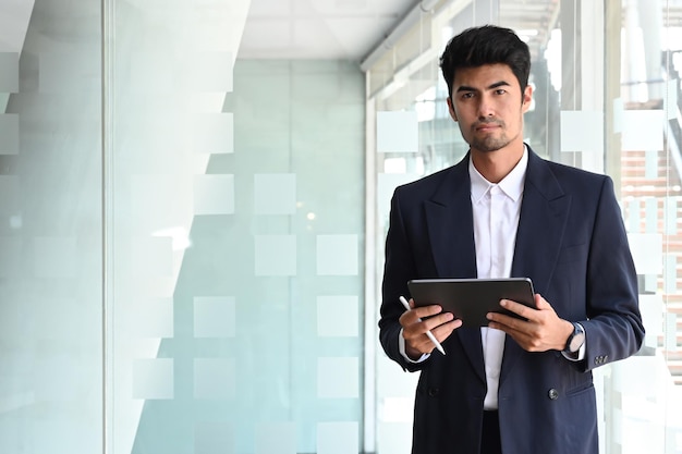 Professioneller Geschäftsmann im schwarzen Anzug mit digitalem Tablet und selbstbewusstem Blick in die Kamera
