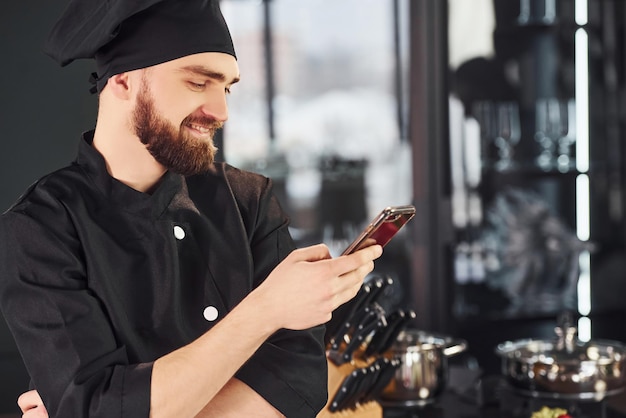 Professioneller junger Koch in Uniform, der mit Telefon in der Küche steht