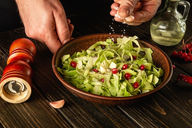 Professioneller Koch streut Salz auf Salat aus frischem Gemüse auf Holztisch