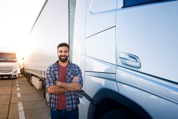 Professioneller LKW-Fahrer mit verschränkten Armen, der durch Sattelzugfahrzeug steht.