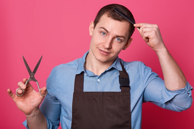 Professioneller männlicher Friseur hält eine Schere in der Hand, kämmt sich, trägt ein blaues Hemd und eine braune Schürze und arbeitet im Friseursalon oder Schönheitssalon