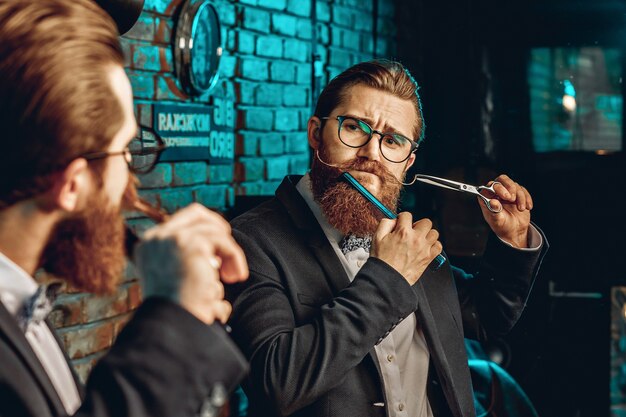 professioneller männlicher Friseur in Gläsern, mit Blick in den Spiegel und einem Kamm und einer Schere neben seinem Gesicht.