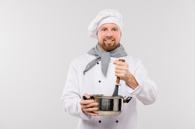 Professioneller männlicher Koch, der Suppe oder andere Mahlzeit in der Pfanne mischt, während Essen vor der Kamera isoliert kocht