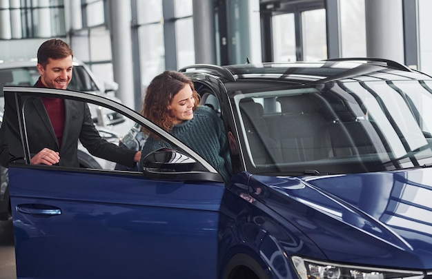 Professioneller Verkäufer, der jungen Mädchen bei der Auswahl neuer moderner Automobile im Innenbereich hilft.