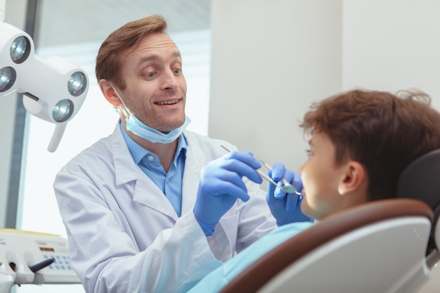 Professioneller Zahnarzt, der freudig mit seinem kleinen Patienten spricht, während er seine Zähne untersucht