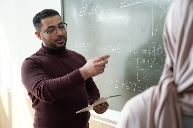 Professor adulto mestiço explicando o assunto ao aluno em hijab no quadro-negro