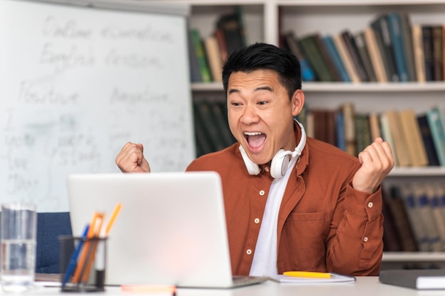 Professor asiático emocional usando laptop gesticulando sim sentado na sala de aula