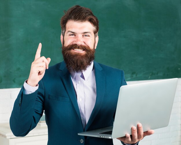 Professor com notebook portátil na sala de aula na escola jovem professor masculino com o dedo apontando perto