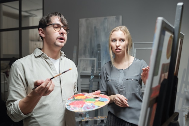 Professor confiante com pincel e paleta e seu aluno discutindo detalhes de uma pintura inacabada no cavalete na aula