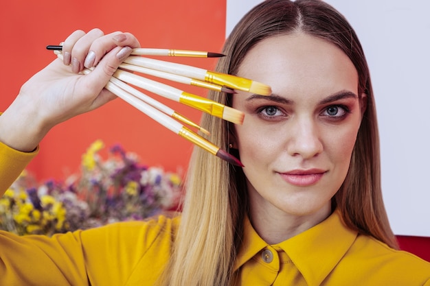 Professor de artes. Uma linda professora loira da escola de arte segurando pincéis perto do rosto