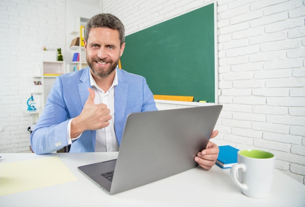 Professor de escola sorridente em sala de aula com computador no quadro-negro