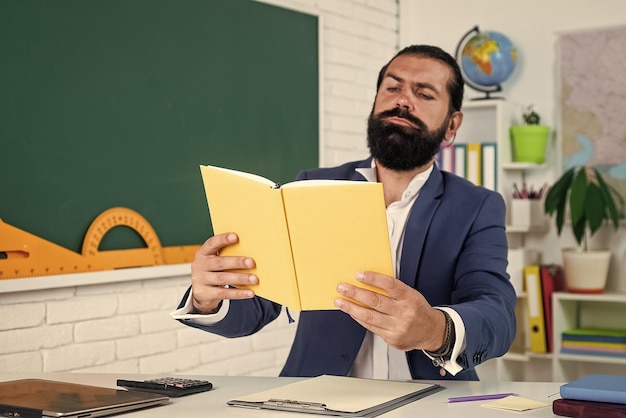 Foto professor de homem barbudo maduro em traje sente-se na sala de aula da escola com quadro-negro ler literatura de livro
