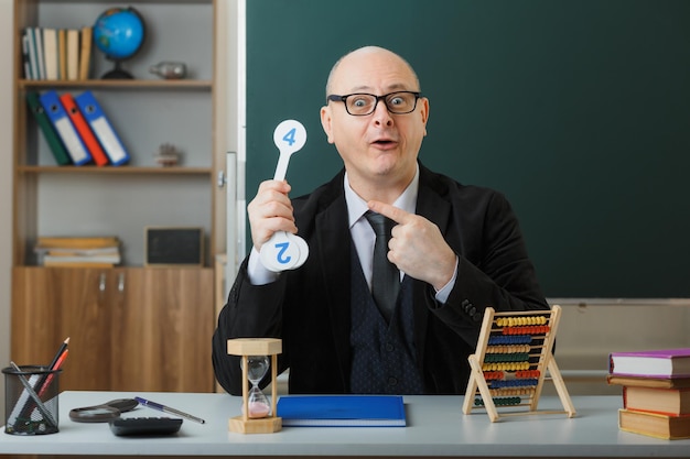 Professor de homem usando óculos sentado na mesa da escola com registro de classe na frente do quadro-negro na sala de aula segurando placas explicando lição apontando com o dedo indicador parecendo surpreso