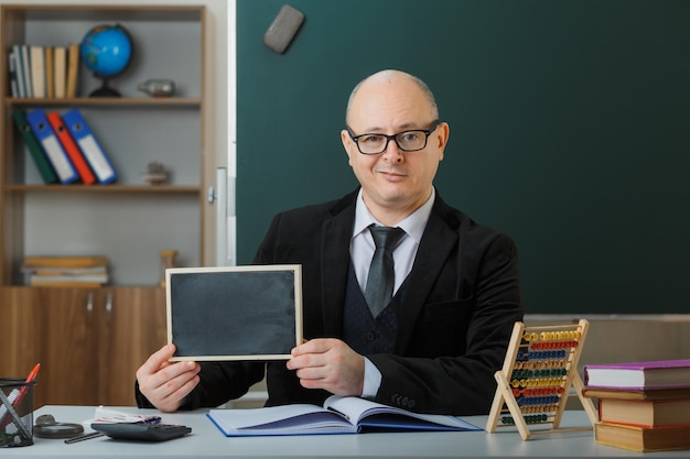 Professor de homem usando óculos sentado na mesa da escola na frente do quadro-negro em sala de aula mostrando lousa explicando lição explicando lição com sorriso no rosto