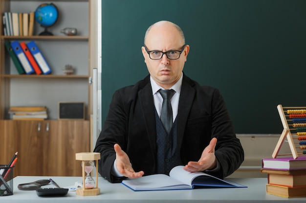 Professor de homem usando óculos verificando o registro de classe olhando para a câmera sendo confuso e descontente sentado na mesa da escola na frente do quadro-negro na sala de aula