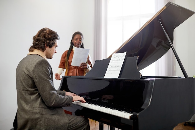 Professor de música jovem tocando piano enquanto seu aluno canta música em pé atrás do piano durante a aula de música