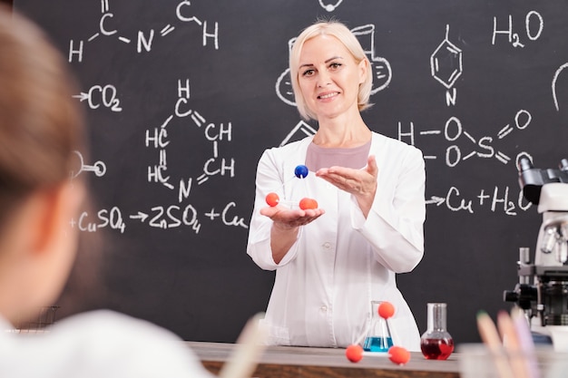 Professor de química sorridente apresentando modelo de molécula de composto químico para crianças em idade escolar no quadro-negro com fórmula química