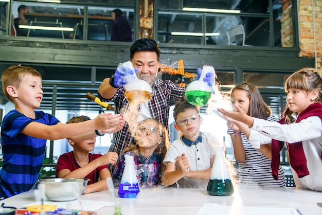 Foto professor jovem asiático com um grupo de seis alunos caucasianos alegres de 8 a 10 anos de óculos protetores durante experimento químico com líquidos coloridos na escola moderna.