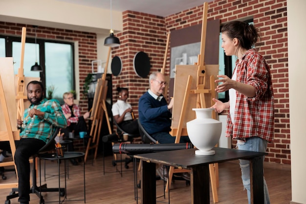 Professor pintor supervisionando homem sênior enquanto desenha modelo de vaso na tela trabalhando na técnica de ilustração no estúdio de criatividade. Equipe diversificada participando da aula de arte desenvolvendo habilidade artística