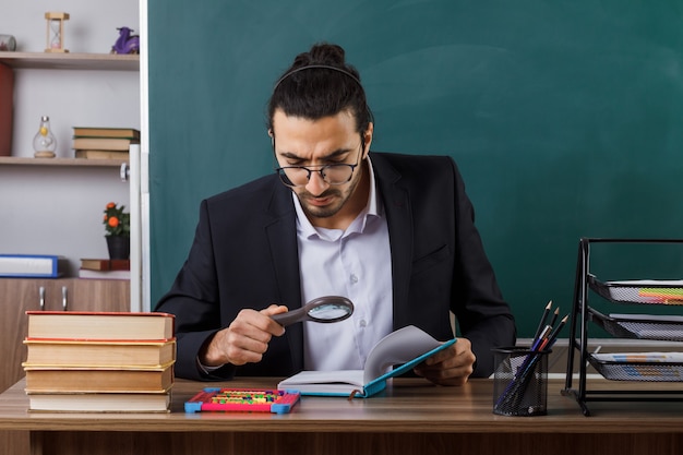 Professor rigoroso de óculos, segurando e lendo um livro com lupa, sentado à mesa com as ferramentas da escola na sala de aula