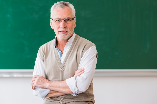 Professor sênior, com, braços cruzaram, olhando câmera, contra, vazio, chalkboard