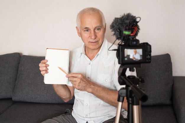 Professor sênior mostrando algo no caderno fazendo um vídeo para palestra em casa