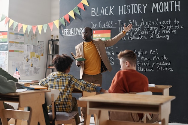 Foto professora afro-americana apontando para o quadro preto e falando sobre o mês da história negra para as crianças da escola durante uma lição