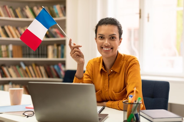 Professora alegre de raça mista dando aula de francês se comunicando no laptop sentado no interior da biblioteca