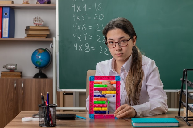 professora de óculos sentada na mesa da escola com contas, preparando a aula, parecendo confiante em frente ao quadro-negro na sala de aula