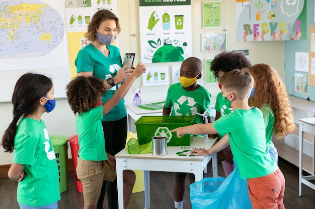 Foto professora e alunos usando máscaras faciais com recipiente de reciclagem na mesa da aula