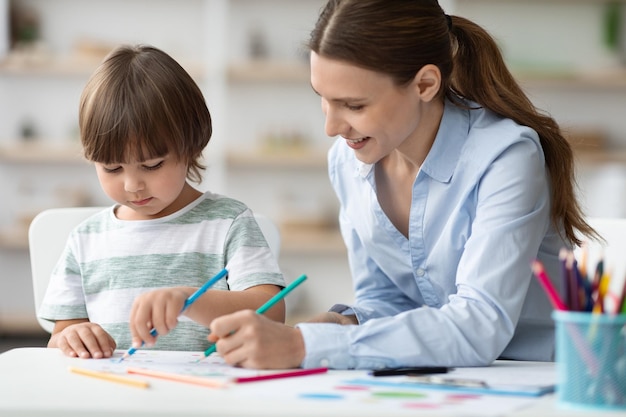 Professora feliz se exercitando com um pequeno estudante menino feliz colorindo fotos apreciando o desenvolvimento