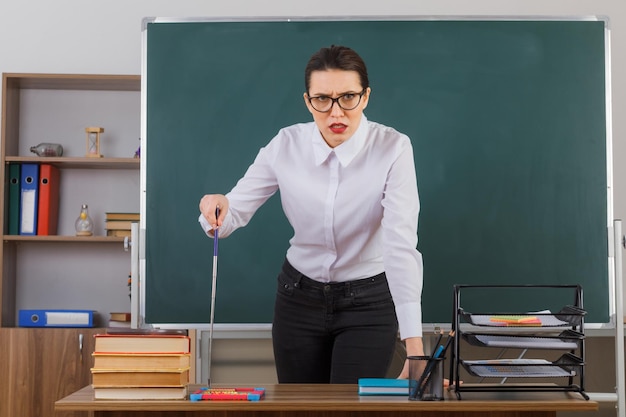 Professora jovem de óculos segurando o ponteiro enquanto explica a lição com expressão séria em pé na mesa da escola na frente do quadro-negro na sala de aula