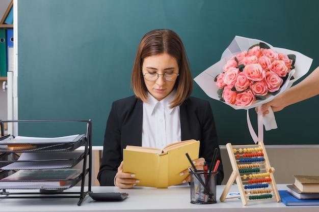 Professora jovem de óculos sentado na mesa da escola na frente do quadro-negro na sala de aula lendo livro preparando-se para a aula ao receber buquê de flores dos alunos