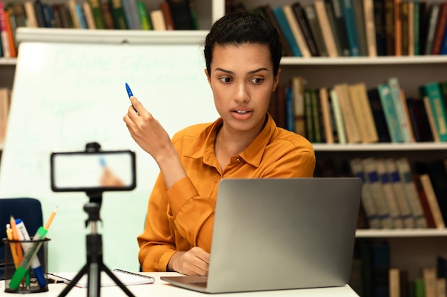 Professora jovem de raça mista em óculos conversando com a webcam do laptop e explicando novo material para os alunos