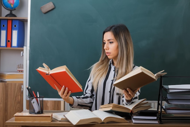 professora jovem sentada na mesa da escola na frente do quadro-negro na sala de aula segurando livros parecendo confuso tentando fazer uma escolha