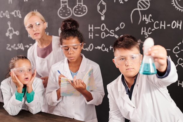 Professora loira e duas colegiais interculturais observando a reação química da mistura de substâncias em um tubo segurado por sua colega de classe