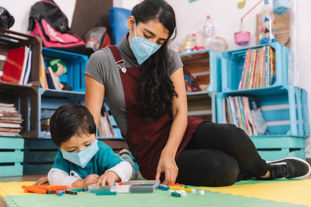 professora mexicana com máscara facial cuidando e brincando com bebê com máscara facial dentro da escola