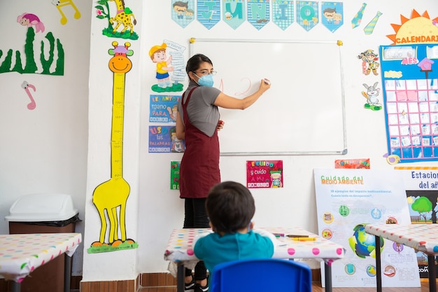 Professora mexicana com máscara facial escrevendo no quadro branco ensinando bebê mexicano na escola